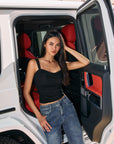 Model wearing BTTRMMT The Brielle Top, black ruched tank with wide straps, styled with blue jeans while standing by a white car.