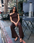 Model wearing the BTTRMMT Daniela Off-Duty Set in brown, seated outdoors in a one-shoulder ruched top and wide-leg pants.