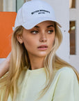 BTTRMMT white Better Moments cap styled on a blonde model in a soft pastel top for a clean casual streetwear look.