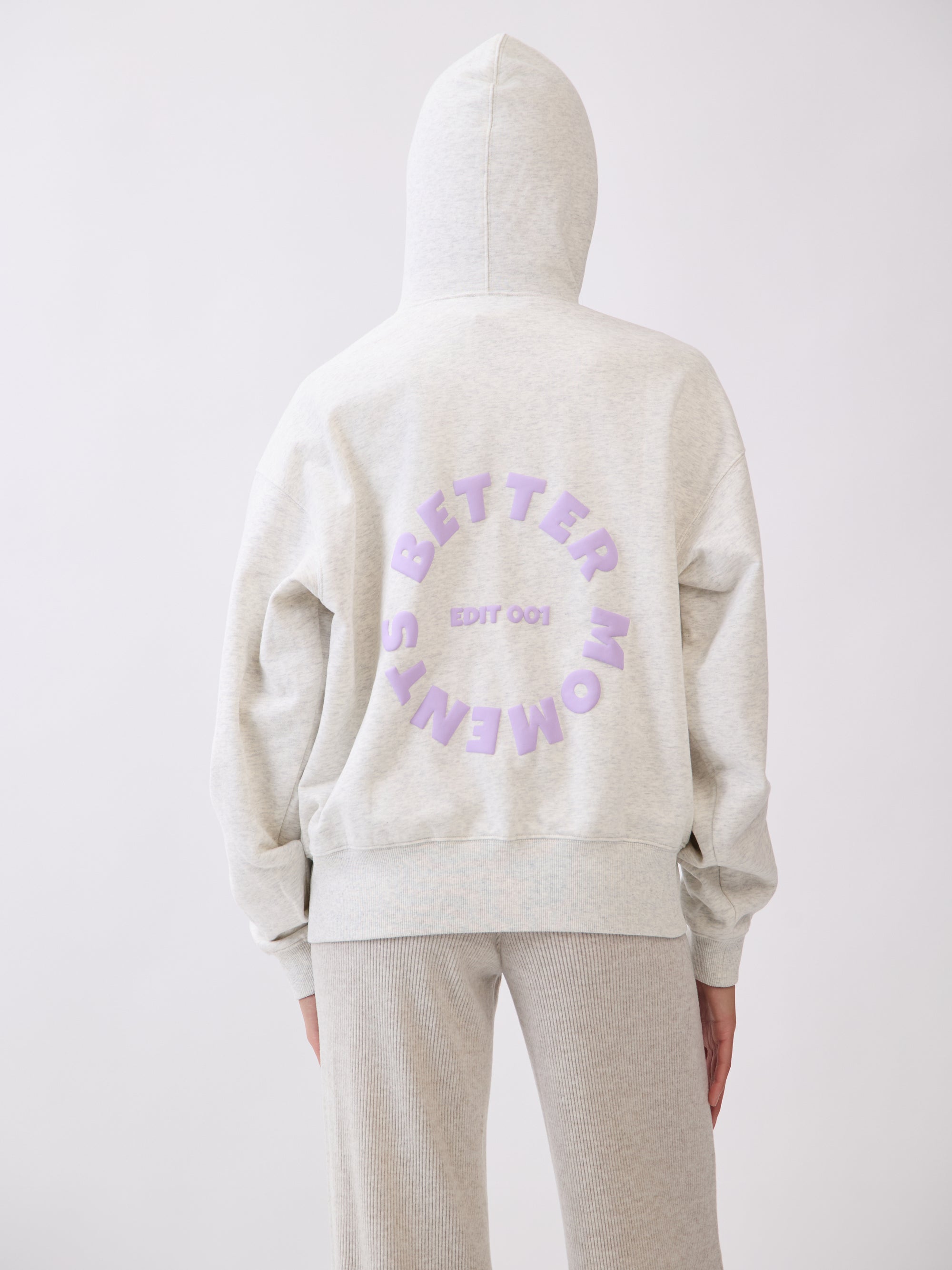 Model wearing BTTRMMT Chill Zip hoodie with lilac “Better Moments Edit 001” logo on the back, cozy minimal streetwear look.