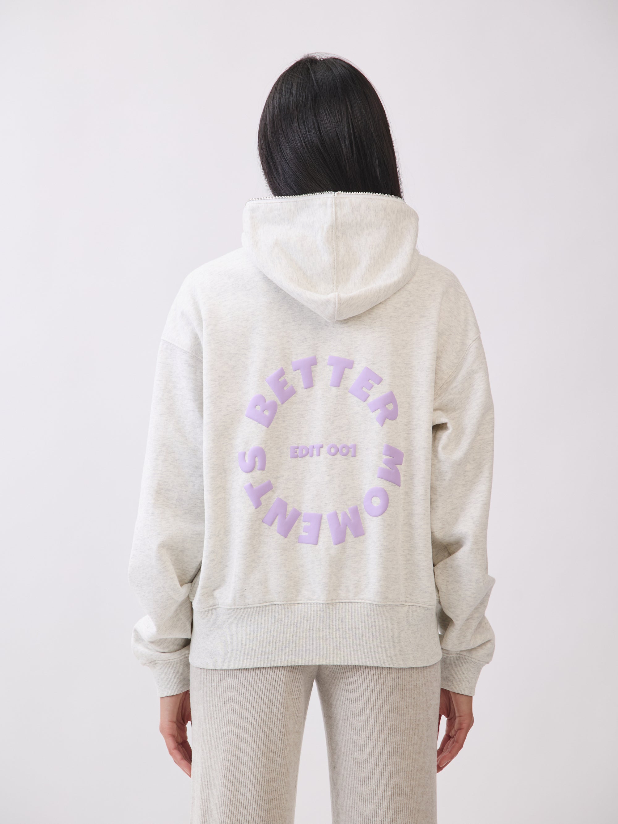 Model wearing BTTRMMT gray hoodie with lilac “Better Moments Edit 001” logo on the back, cozy minimal streetwear look.