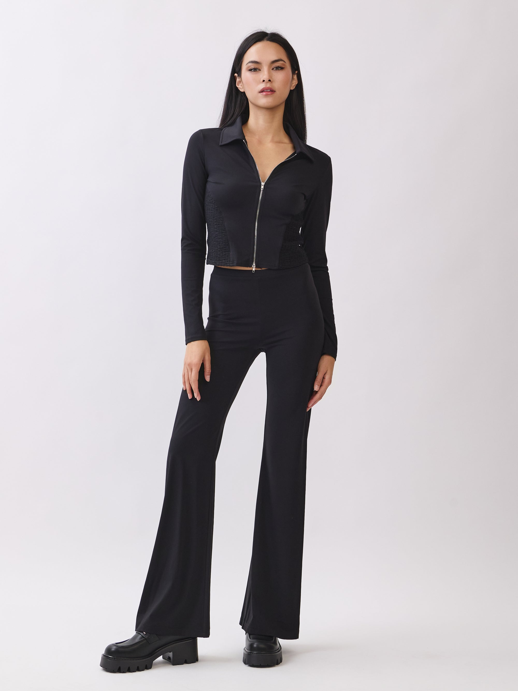 Model wearing BTTRMMT Calista Fit Flare Pants in black with a smooth waistband and flared legs, styled with a fitted black zip-up top.