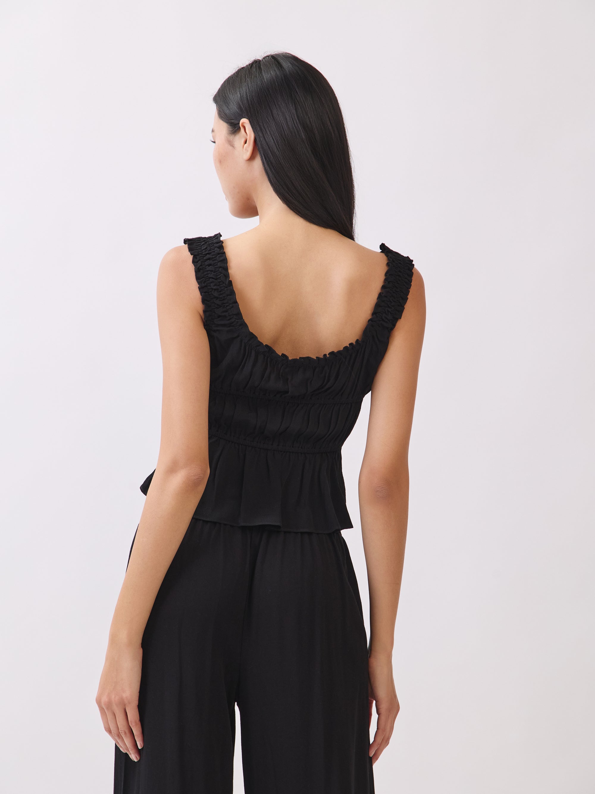 Back view of BTTRMMT black ruffle sleeveless top showing gathered detail, styled with black pants for a clean minimal look