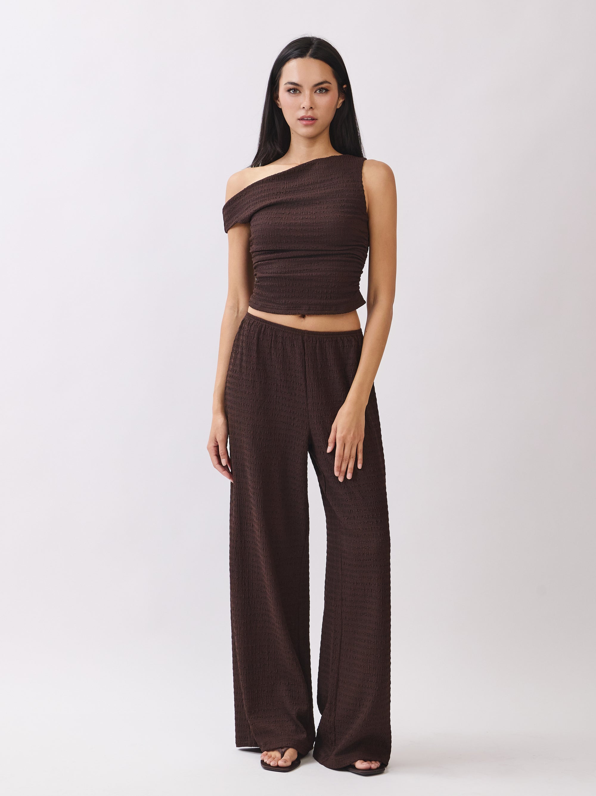 Model wearing the BTTRMMT Daniela Off-Duty Set in brown, featuring a one-shoulder ruched top and matching wide-leg pants for an elevated casual look.