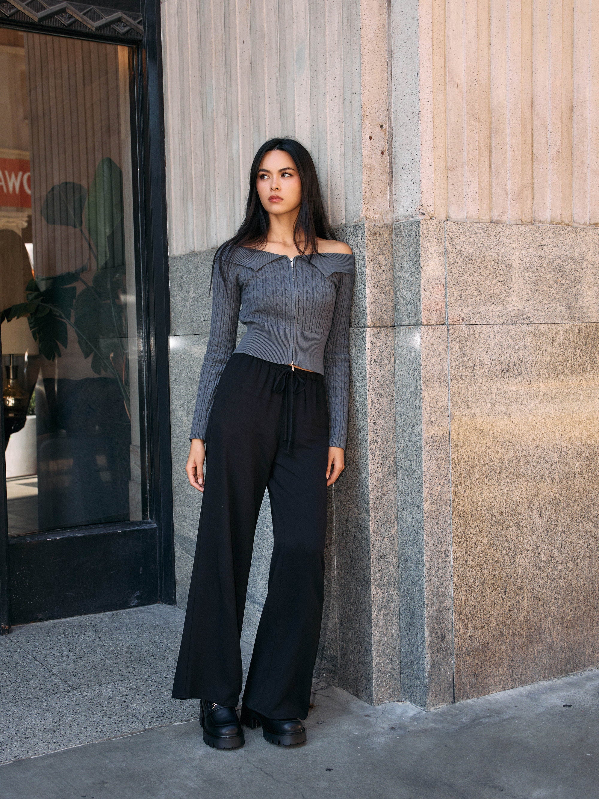 Model wearing the BTTRMMT Camille Zip Knit in charcoal gray with black wide-leg pants, styled outdoors against a concrete building.
