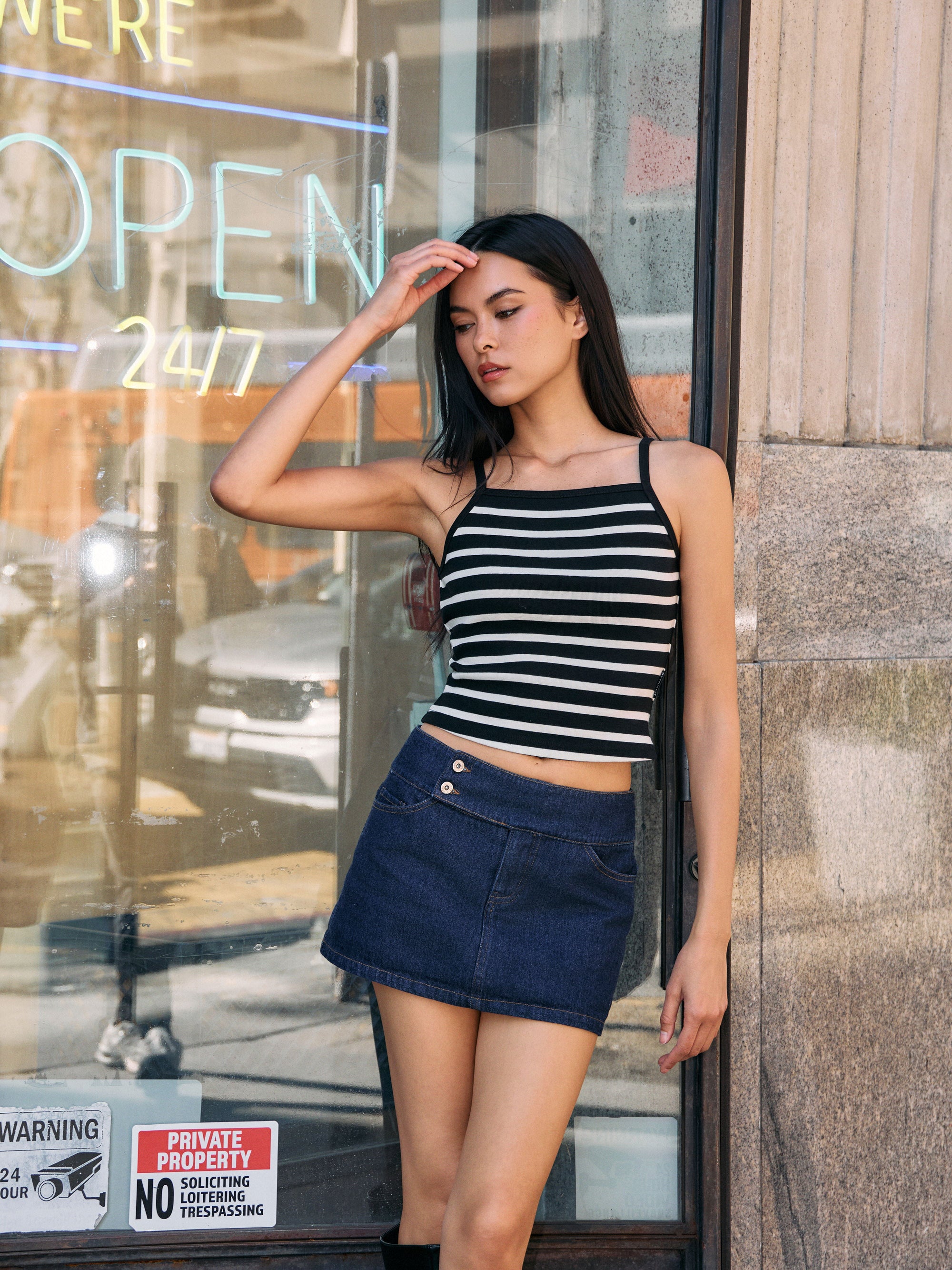 Model wearing ivory and black striped ribbed tank top with denim mini skirt, standing outdoors by glass storefront.