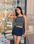Model wearing ivory and black striped ribbed tank top with denim mini skirt, standing outdoors by glass storefront.