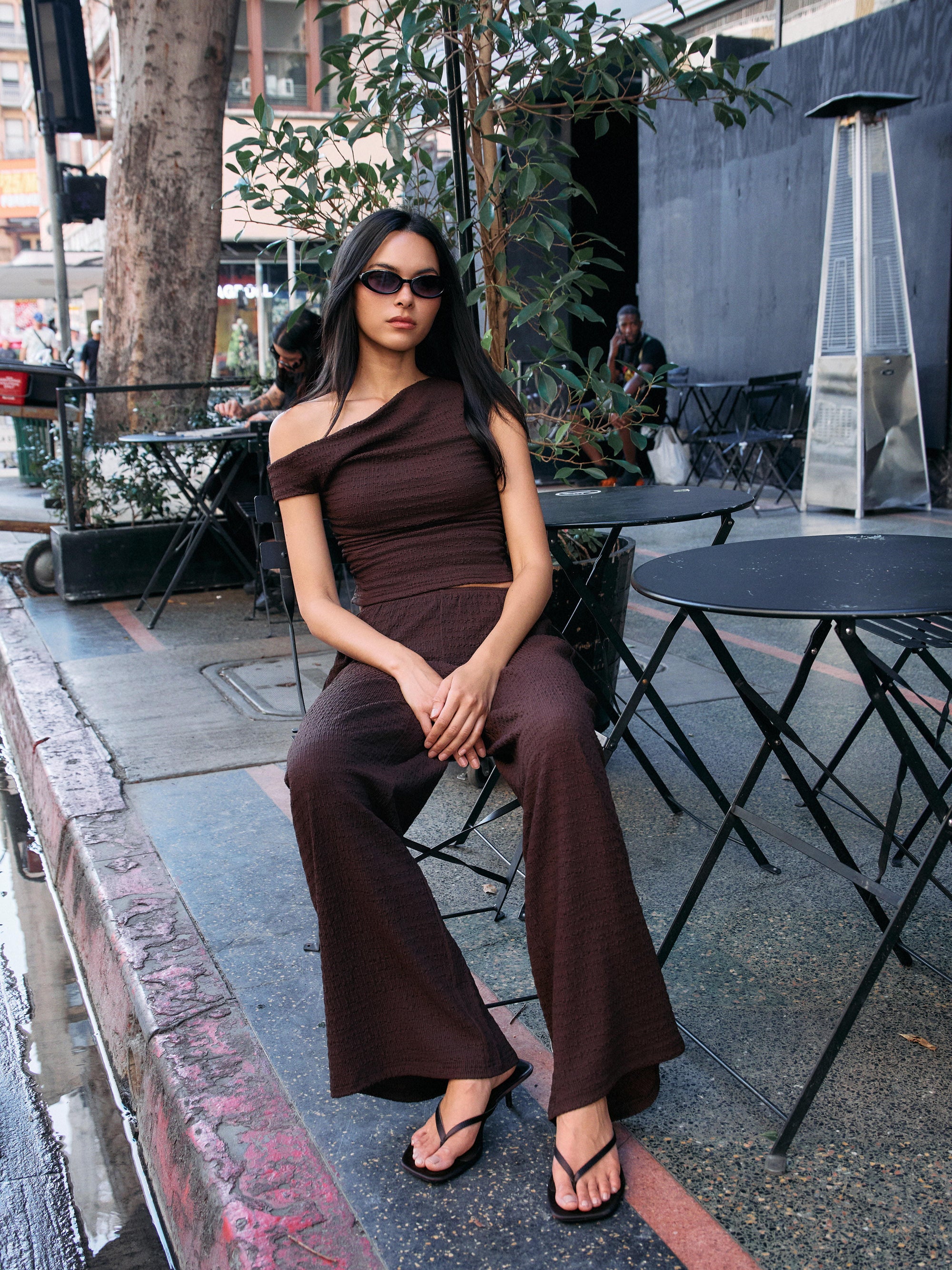 Model wearing the BTTRMMT Daniela Off-Duty Set in brown, seated outdoors in a one-shoulder ruched top and wide-leg pants.