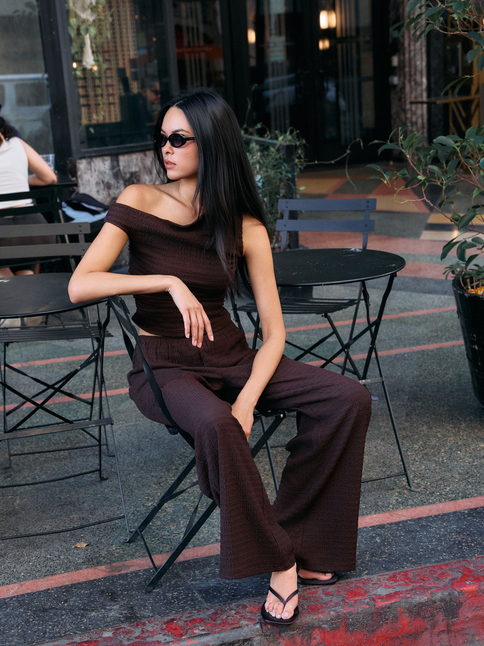 Model wearing the BTTRMMT Daniela Off-Duty Set in brown, seated outdoors in a one-shoulder ruched top and wide-leg pants.
