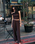 Model wearing the BTTRMMT Daniela Off-Duty Set in brown, seated outdoors in a one-shoulder ruched top and wide-leg pants.